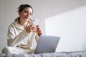 bellissimo sorridente donna adolescente ragazza alunno libero professionista con buio lungo capelli nel casuale utilizzando mobile Telefono Lavorando su il computer portatile seduta su divano a il casa foto