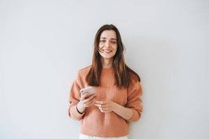 ritratto di bellissimo sorridente donna alunno con buio lungo capelli nel casuale a maglia maglione utilizzando mobile Telefono nel mano su il grigio sfondo foto