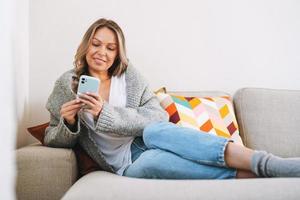 giovane bellissimo donna quaranta anno con bionda lungo Riccio capelli nel accogliente a maglia grigio maglione con mobile Telefono nel mani seduta su divano a casa foto