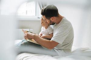contento padre giovane uomo e bambino ragazza poco figlia avendo divertimento lettura un' libro nel bambini camera a casa foto