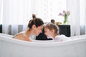 giovane madre donna con lungo capelli con poco gemello ragazza figlia nel pigiama avendo divertimento nel il bagno con schiuma a casa foto