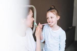 giovane madre donna con lungo capelli con poco gemello ragazza figlia nel pigiama spazzolatura loro denti nel il mattina a casa foto