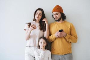 giovane famiglia con bambino poco ragazza su grigio parete sfondo, genitori utilizzando mobile telefoni foto