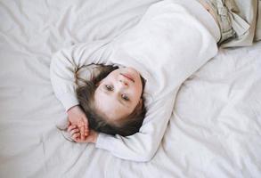 carino divertente poco ragazza con lungo capelli nel bianca casa vestito avendo divertimento bugie su letto a il casa, Visualizza a partire dal superiore foto