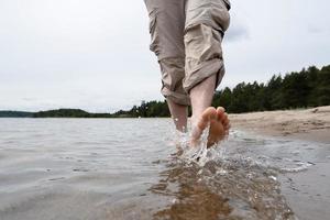 a piedi nudi maschio gambe nel arrotolato pantaloni camminare su il acqua lungo il sabbioso riva contro il fondale di il lago e alberi. stile di vita. foto