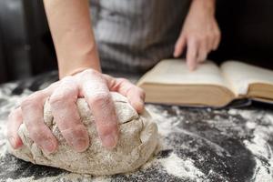 panettiere detiene uno mano su il Impasto e il altro su il libro di cucina, su il tavolo cosparso con Farina. pane fabbricazione. avvicinamento. foto