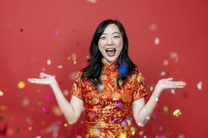 asiatico donna con rosso cheongsam o qipao eccitante e ridendo per desiderando il bene fortuna e prosperità nel Cinese nuovo anno con coriandoli celebrazione vacanza isolato su rosso sfondo concetto foto