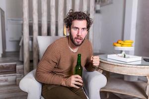 eccitato giovane uomo applauso mentre Guardando gli sport incontro su divano a casa. attraente, contento tipo Guardando calcio, festeggiare vittoria di il suo preferito squadra, urlando, avendo birra . foto