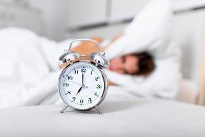 frustrato donna copertura sua orecchie con sua cuscino a partire dal allarme orologio nel il mattina. ritratto di infastidito giovane donna copertura orecchie con cuscino dovuto per suono allarme orologio nel mattina, mentre dire bugie nel letto foto