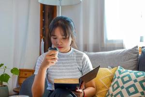 asiatico donna sorseggiando caffè e leggere un' libro su il vivente camera divano nel loro gratuito volta. concetto rilassare dopo opera, relax, accogliente angolo, calma angolo. morbido e selettivo messa a fuoco. foto