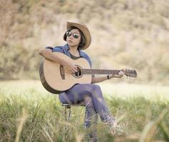 donne corto capelli indossare cappello e occhiali da sole sedersi giocando chitarra nel erba campo foto