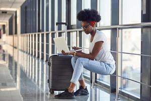 giovane africano americano femmina passanger nel casuale Abiti è nel aeroporto con Bagaglio e utilizzando il computer portatile foto