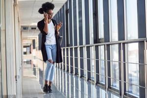 giovane africano americano femmina passanger nel casuale Abiti è nel aeroporto ascoltando musica nel cuffie foto