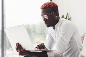 giovane africano americano uomo nel formale Abiti seduta in casa con il computer portatile nel mani foto