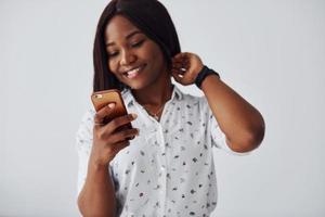 positivo africano americano donna nel bianca camicia in piedi in casa contro bianca parete con Telefono nel mano foto