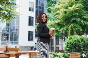 caffè rompere. giovane afro americano donna nel alla moda Abiti all'aperto nel il città vicino verde alberi e contro attività commerciale edificio foto
