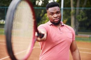 africano americano uomo nel rosa camicia in posa per un' telecamera con tennis racchetta su il Tribunale all'aperto foto