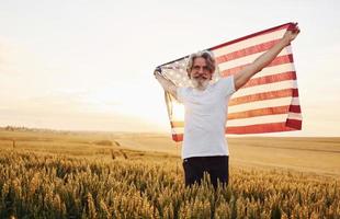 Tenere Stati Uniti d'America bandiera nel mani. patriottico anziano elegante uomo con grigio capelli e barba su il agricolo campo foto