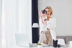 di mezza età donna nel elegante Abiti è a casa. fotografo con telecamera foto