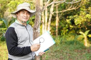 bello asiatico uomo botanico è a foresta per sondaggio botanico impianti, detiene carta appunti e ingrandimento bicchiere. concetto, esaminare, Esplorare, ricerca, studia di ambiente, impianti e natura. foto