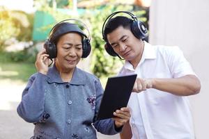 asiatico anziano donna e asiatico uomo indossa cuffia, detiene inteligente tavoletta all'aperto. concetto, figlio dà consigli mamma Come per uso in linea media a partire dal inteligente tavoletta. apprendimento nuovo cosa a partire dal Internet. foto