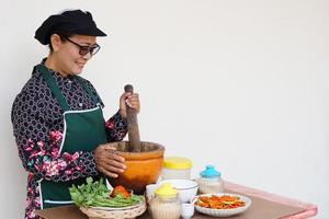 contento asiatico anziano donna è cucinando, indossare capocuoco berretto e grembiule, detiene pestello, mortaio e piatto di peperoncini. concetto, cucinando per famiglia. tailandese cucina stile di vita. anziano attività. foto