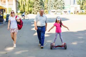 poco sorridente ragazza apprendimento per cavalcata un' hoverboard con sua genitori all'aperto foto