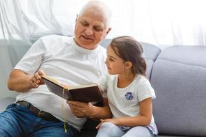 contento poco ragazza con nonno lettura storia libro a casa foto