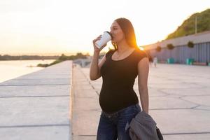 giovane incinta donna potabile porta via caffè o tè su argine. maternità, tempo libero e Salute concetto. pubblicità la zona spazio di lavoro foto