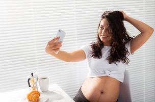 incinta donna prende autoscatto su sua smartphone mentre prima colazione. maternità partire e tecnologie concetto. foto