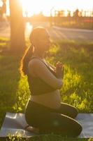 tranquillo, calmo giovane positivo incinta donna nel ginnastica completo da uomo fa yoga e meditare seduta su stuoia su verde erba su soleggiato caldo estate giorno. concetto di preparazione per parto e positivo atteggiamento foto