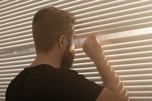 posteriore Visualizza di giovane uomo con barba sbircia attraverso buco nel il finestra persiane e sembra su in il strada. sorveglianza e curiosità concetto foto