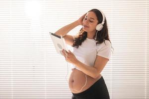 bellissimo giovane incinta donna sceglie musica di sottoscrizione e ascolta per esso attraverso cuffie e tavoletta nel il mattina. concetto di benefici per fetale sviluppo foto