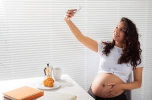 incinta donna prende autoscatto su sua smartphone mentre prima colazione. maternità partire e tecnologie concetto. foto