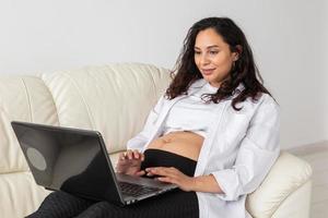 latino incinta donna utilizzando il computer portatile seduta su divano a casa. gravidanza e informazione per paternità concetto. foto