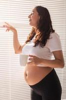 curioso giovane bellissimo incinta donna potabile tè e guardare attraverso il persiane a il finestra. concetto di gioia e bene notizia mentre in attesa per bambino foto