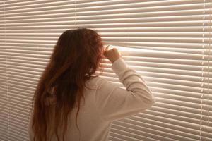 posteriore Visualizza di bellissimo giovane donna con lungo capelli sbircia attraverso buco nel il finestra persiane e sembra su il finestra. sorveglianza e curiosità concetto foto