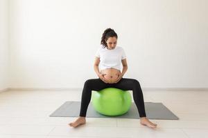 un' giovane incinta donna fare rilassamento esercizio utilizzando un' fitness palla mentre seduta su un' stuoia e Tenere sua pancia. foto