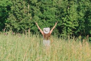 posteriore Visualizza di contento giovane donna con lungo capelli nel cappello e vestito tira sua braccia su mentre a piedi attraverso estate foresta su un' soleggiato giorno. gioia di vita concetto foto