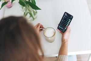 giovane donna controllo criptovaluta grafici e azione citazioni su mobile Telefono con tazza di caffè nel il cucina a casa, Visualizza a partire dal superiore foto