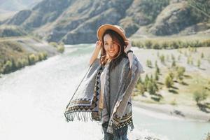 spensierato brunetta giovane donna viaggiatore nel poncho a partire dal indietro su sfondo di il montagna fiume foto