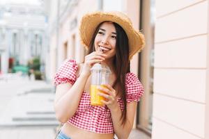 sorridente bellissimo asiatico giovane donna con lungo capelli nel cannuccia cappello potabile arancia succo a città strada foto