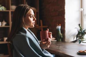 giovane bella donna nel accogliente grigio sciarpa con boccale di tè nel mani sembra su il finestra e riposa nel sua studio foto