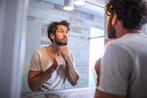 riflessione di bello uomo con barba guardare a specchio e toccante viso nel bagno toelettatura. uomo mettendo cura della pelle facciale trattamento crema su viso. foto