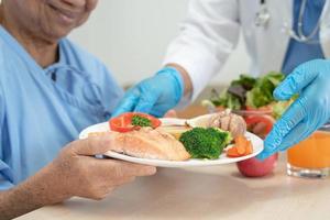 asiatico anziano vecchio signora donna paziente mangiare prima colazione e verdura salutare cibo con speranza e contento mentre seduta e Affamato su letto nel Ospedale. foto