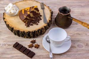 superiore Visualizza di un' pezzo di cioccolato torta su di legno ceppo con un' caffè tazza, tè cucchiaio, forchetta, anice, caffè fagioli, cioccolato bar e ciotola con zucchero cubi su un' luminosa di legno sfondo foto