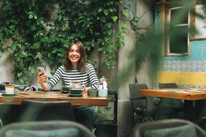 giovane sorridente brunetta donna nel casuale Abiti utilizzando sociale media su mobile Telefono seduta a il verde bar foto