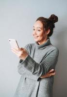 adulto bellissimo sorridente donna quaranta anni con brunetta Riccio capelli nel caldo grigio a maglia vestito utilizzando mobile su grigio sfondo foto