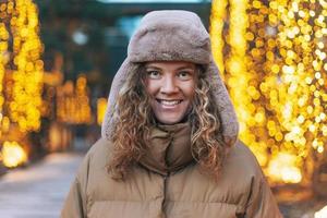ritratto di giovane sorridente donna con Riccio capelli nel pelliccia cappello nel il inverno strada decorato con luci foto