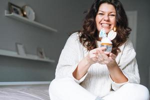 giovane adulto contento bellissimo donna quaranta anni più dimensione corpo positivo nel bianca accogliente a maglia maglione con festivo Cupcake con candela nel mani su letto a il casa festa, compleanno ragazza foto